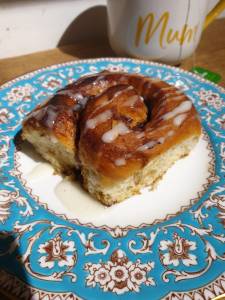 Vegan cinnamon bun drizzled with icing on a blue and gold floral plate with a yellow and white mug behind. 